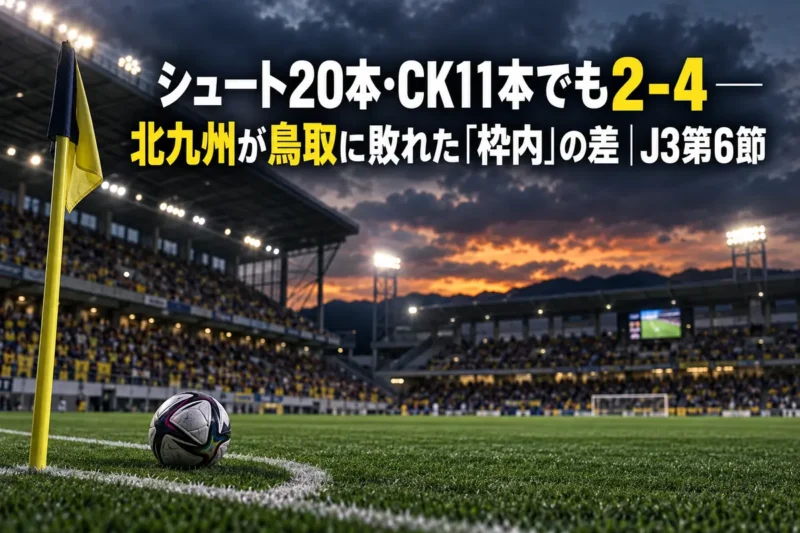 夕方の曇り空の下、照明が灯るサッカースタジアムのピッチとコーナーフラッグ付近に置かれたボール。