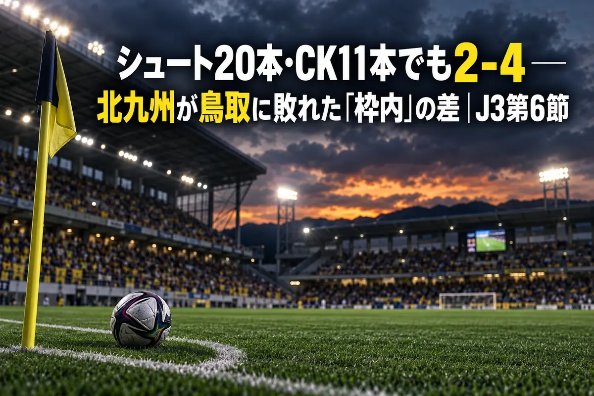 夕方の曇り空の下、照明が灯るサッカースタジアムのピッチとコーナーフラッグ付近に置かれたボール。