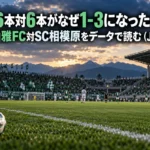 山あいのサッカースタジアムで行われる試合を客席側から捉えた風景写真。