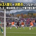 秋の午後のスタジアムで、終盤のゴール前で競り合う両チームの選手と、タッチラインから入る交代選手。